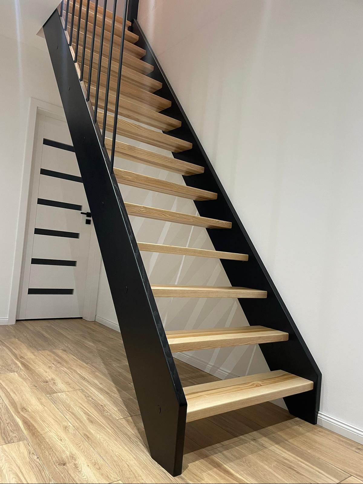 Nowoczesne schody o drewnianych stopniach i czarnej, metalowej konstrukcji. Minimalistyczny design, widok z dołu, drzwi w tle. Jasne drewno, proste linie.