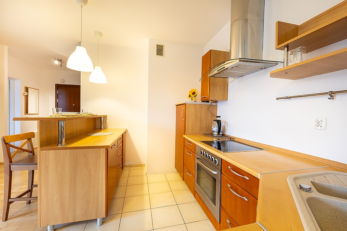 Jasna kuchnia z drewnianymi meblami, blatem i barkiem, z widocznym piekarnikiem, płytą grzewczą, okapem oraz elementami dekoracyjnymi jak słonecznik w wazonie i szklanki na blacie.