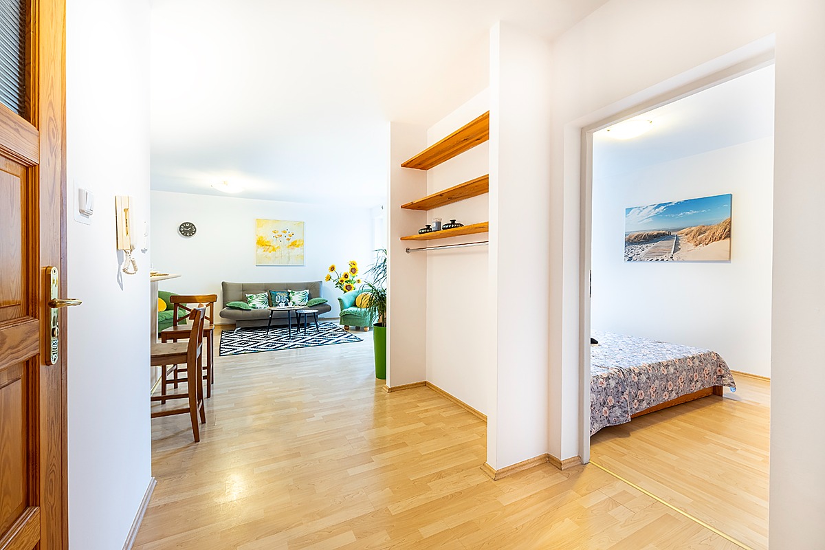 Jasne wnętrze mieszkania z widokiem na salon z szarą sofą, zielonym fotelem i czarno-białym dywanem, oraz sypialnię z łóżkiem i obrazem plaży na ścianie, widoczne z korytarza z drewnianą podłogą...