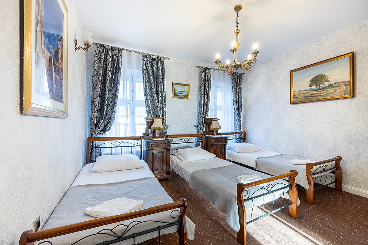 Pokój trzyosobowy z trzema pojedynczymi łóżkami, drewnianymi szafkami nocnymi z lampkami, wzorzystymi tapetami i zasłonami w stylu retro oraz żyrandolem z sześcioma ramionami.