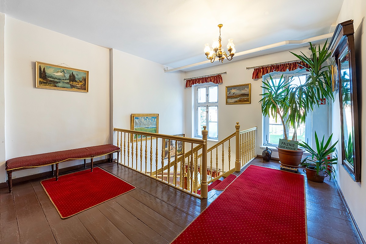 Wnętrze klatki schodowej z drewnianą balustradą, czerwonym dywanem, ławką i obrazami w złotych ramach, oświetlone żyrandolem i naturalnym światłem z okna, z rośliną doniczkową i tabliczką...