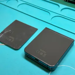 Uszkodzona tylna obudowa składanego smartfona leży obok nowego, sprawnego elementu na niebieskiej macie serwisowej z narzędziami w tle.