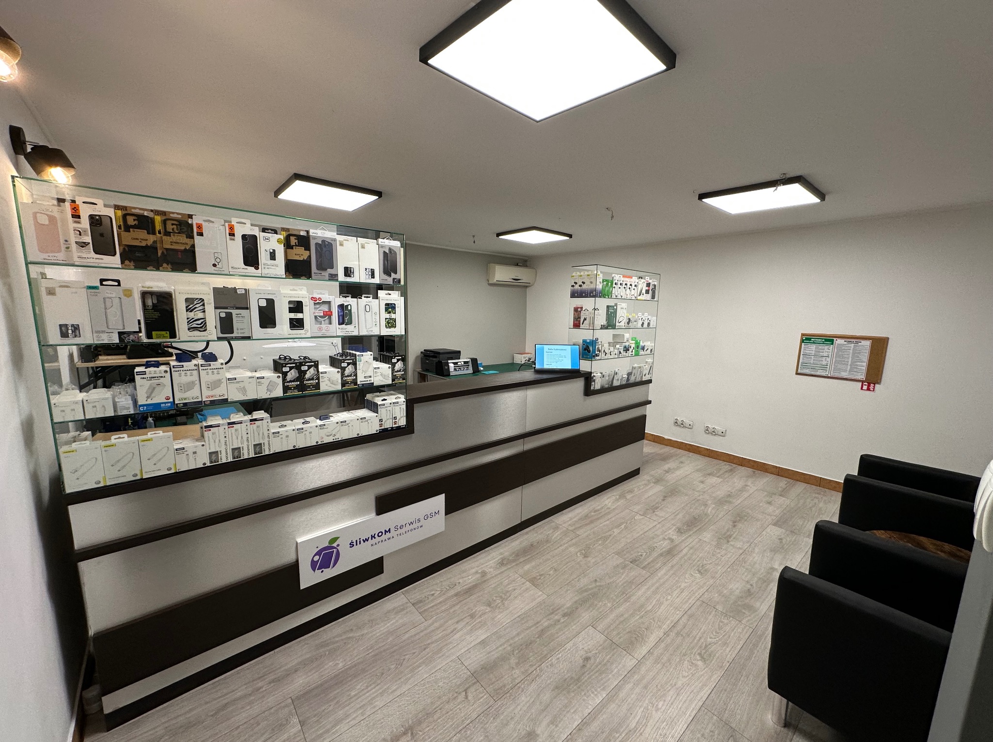Wnętrze punktu usługowego z ladą, za którą znajdują się szklane gabloty z akcesoriami do urządzeń mobilnych, w tle ściana z tablicą informacyjną i krzesła dla klientów.