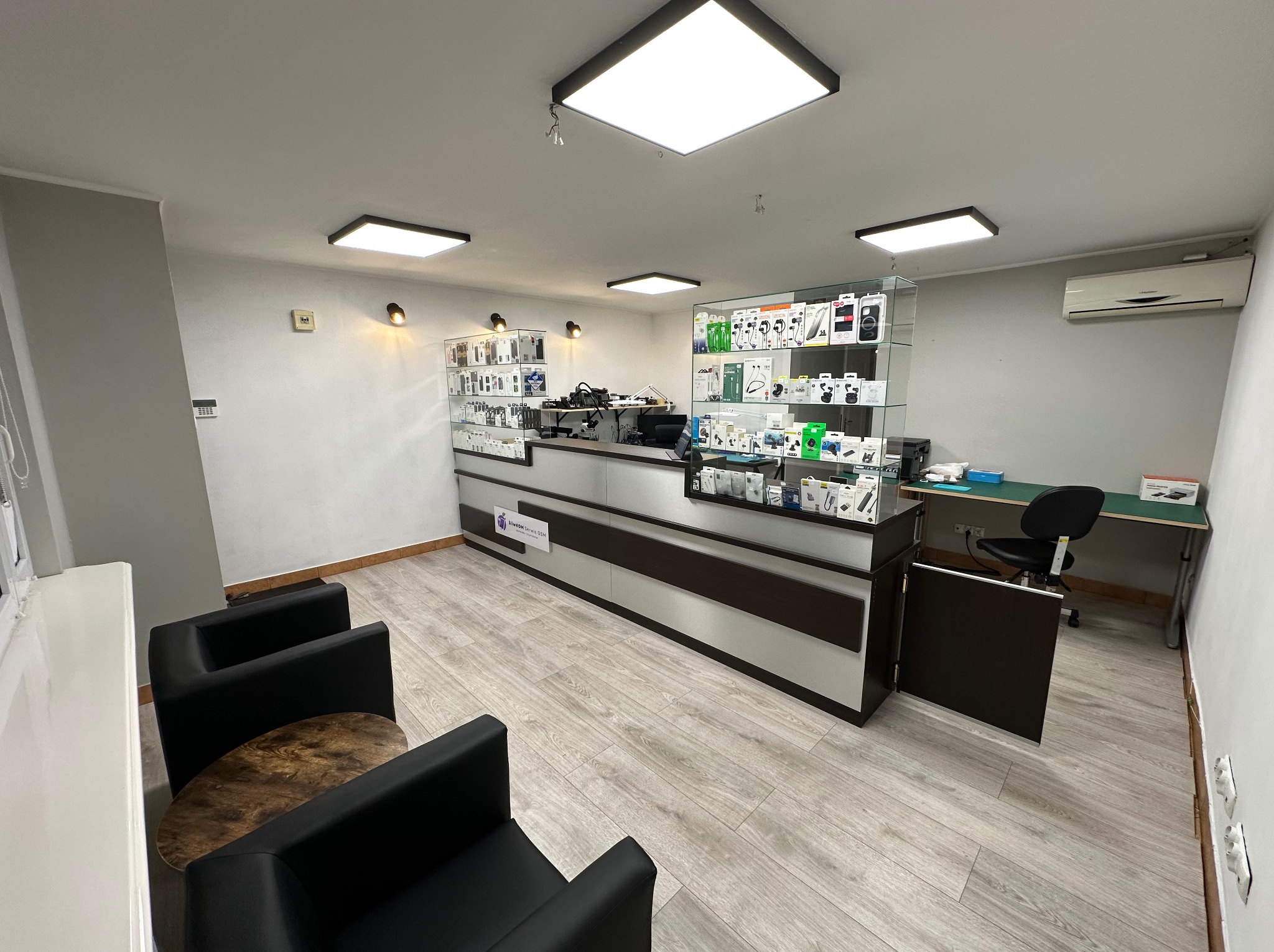 Wnętrze punktu naprawy urządzeń elektronicznych: lada z ekspozycją akcesoriów, stanowisko pracy z krzesłem, dwie witryny z towarami oraz stolik kawowy z fotelami dla klientów.