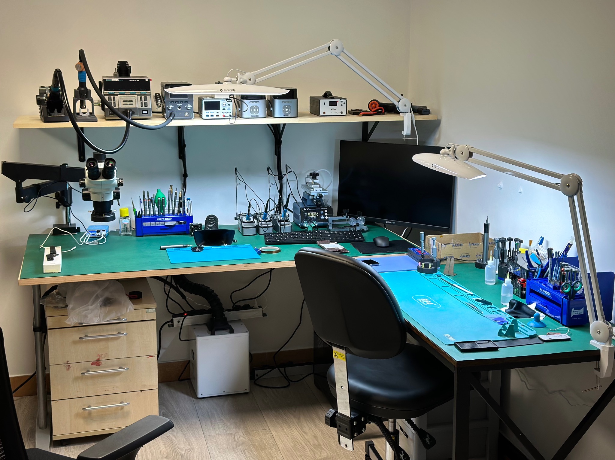 Stanowisko pracy z mikroskopem, lutownicą i narzędziami precyzyjnymi na zielonym blacie, oświetlone lampami, na półce powyżej dodatkowy sprzęt elektroniczny.