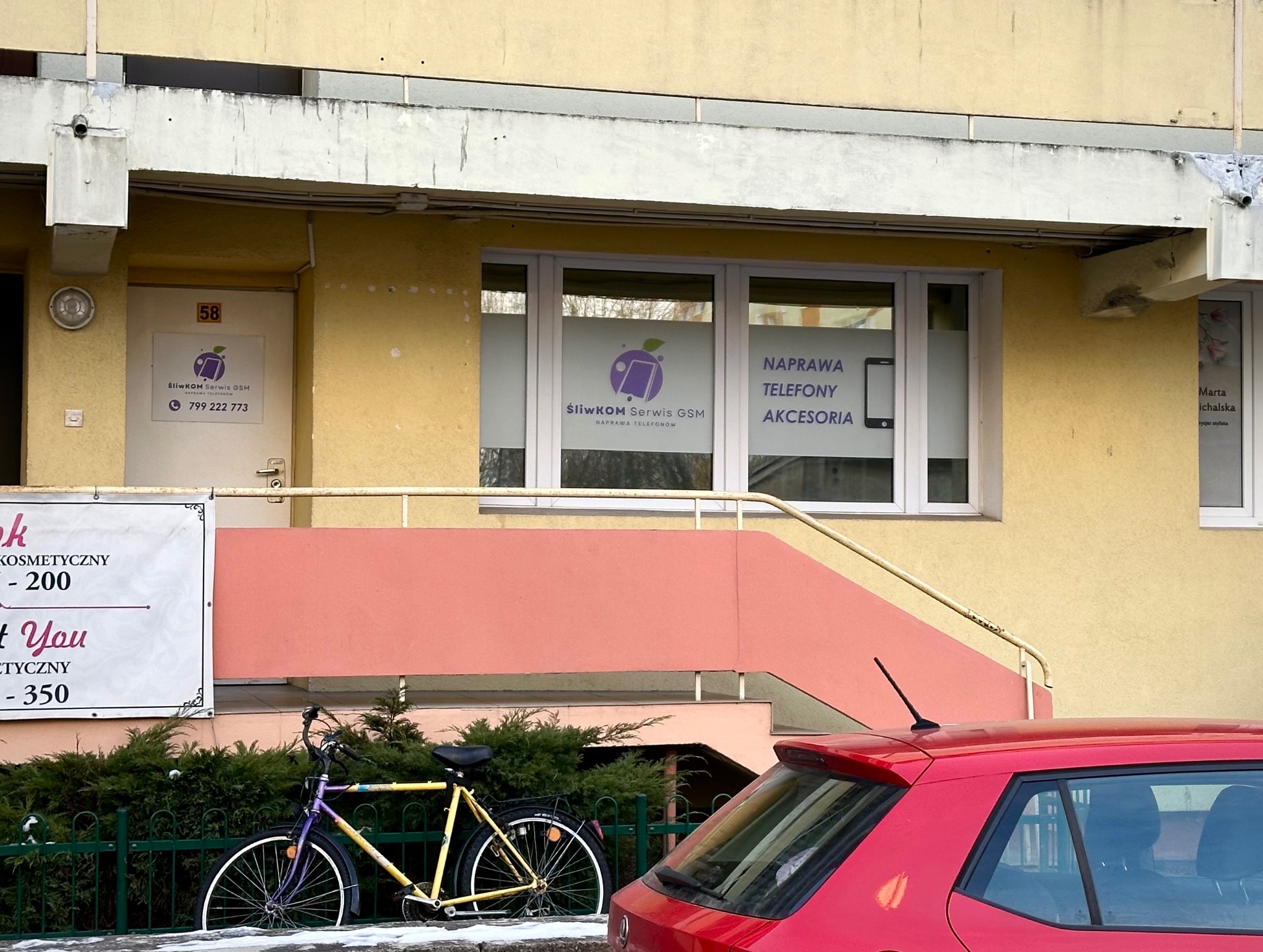 Fasada budynku z szyldem firmy świadczącej usługi GSM, rower oparty o płot i fragment czerwonego samochodu na pierwszym planie.
