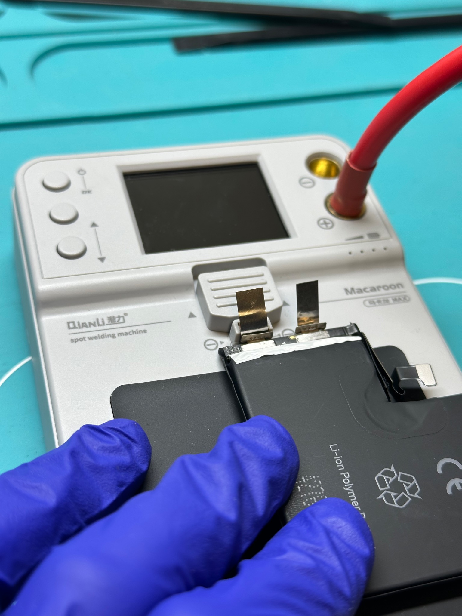 Bateria litowo-polimerowa smartfona podłączona do punktowej spawarki, operator w niebieskich rękawiczkach, widoczne elektrody i wyświetlacz urządzenia.