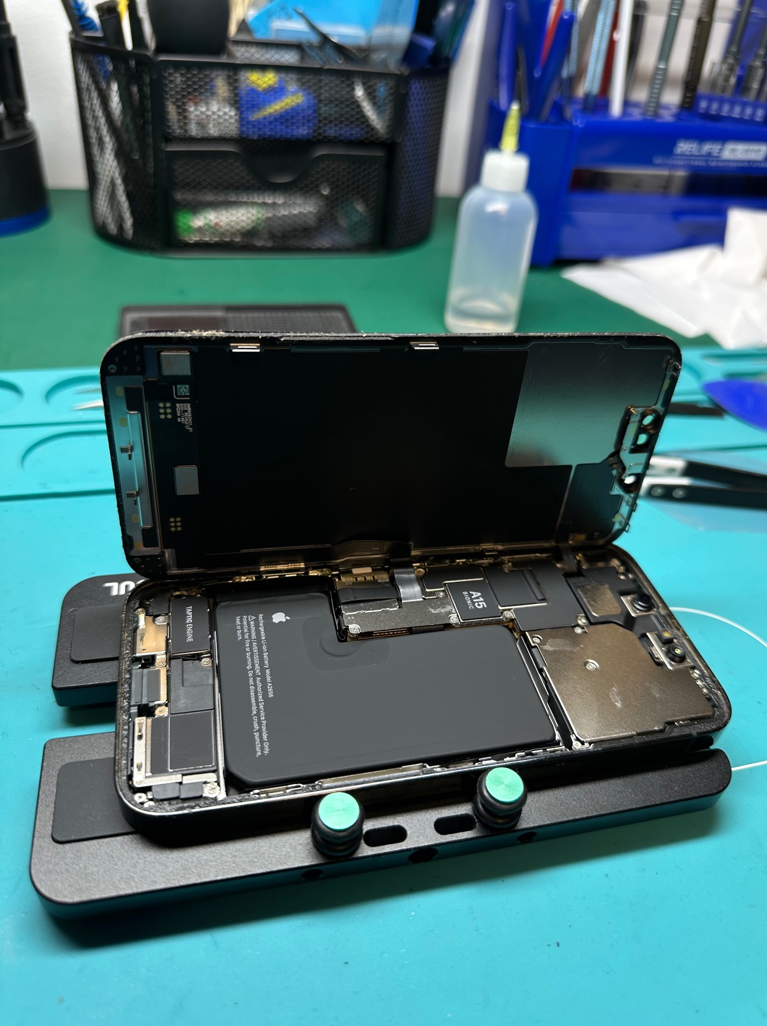 Otwarty smartfon na specjalistycznym uchwycie, widoczne wnętrze z baterią i układami scalonymi, na tle narzędzia i akcesoria serwisowe.