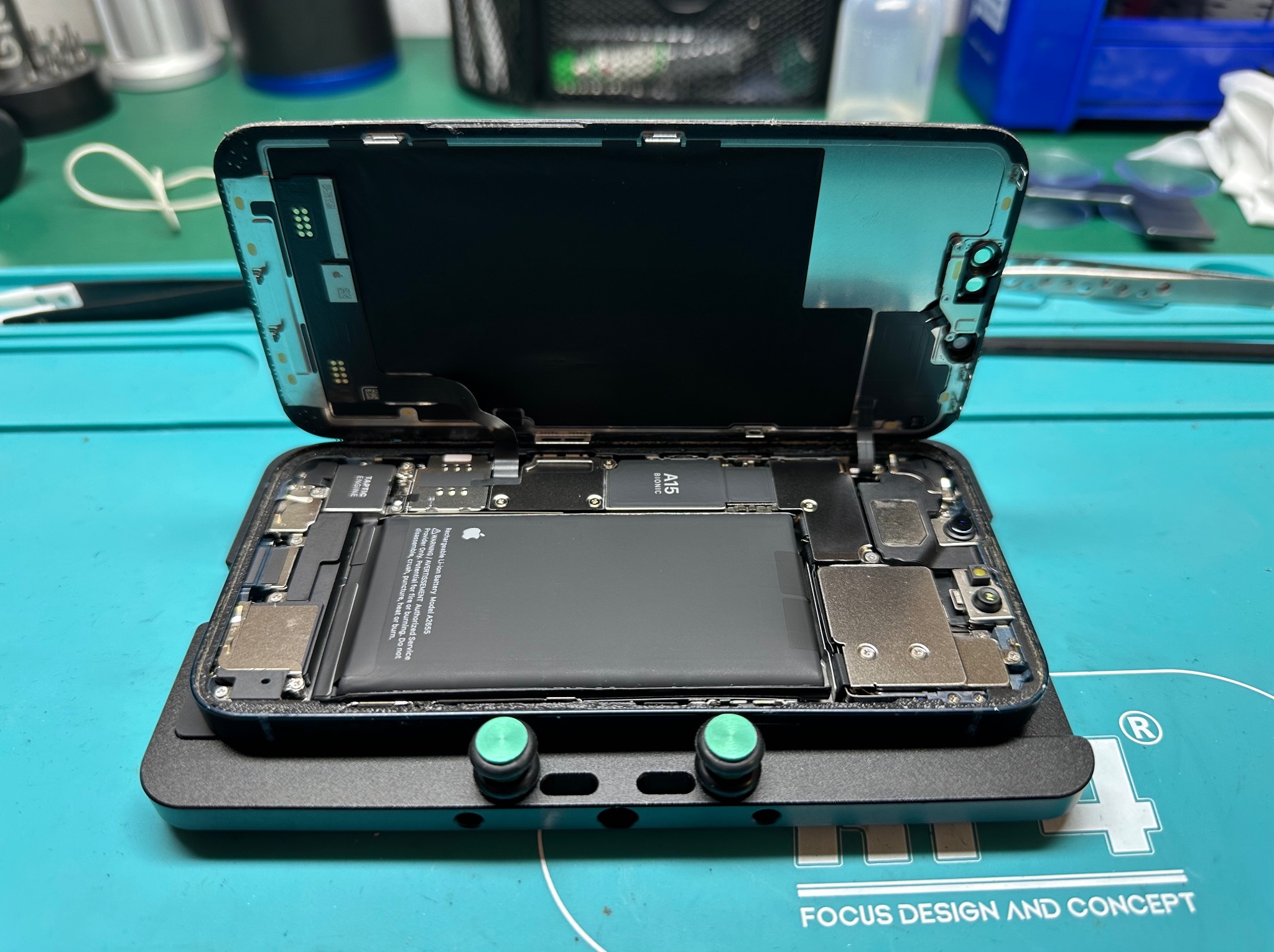 Otwarty iPhone na uchwycie serwisowym, widoczna bateria, płyta główna z chipem A15 Bionic i elementy mocujące ekran