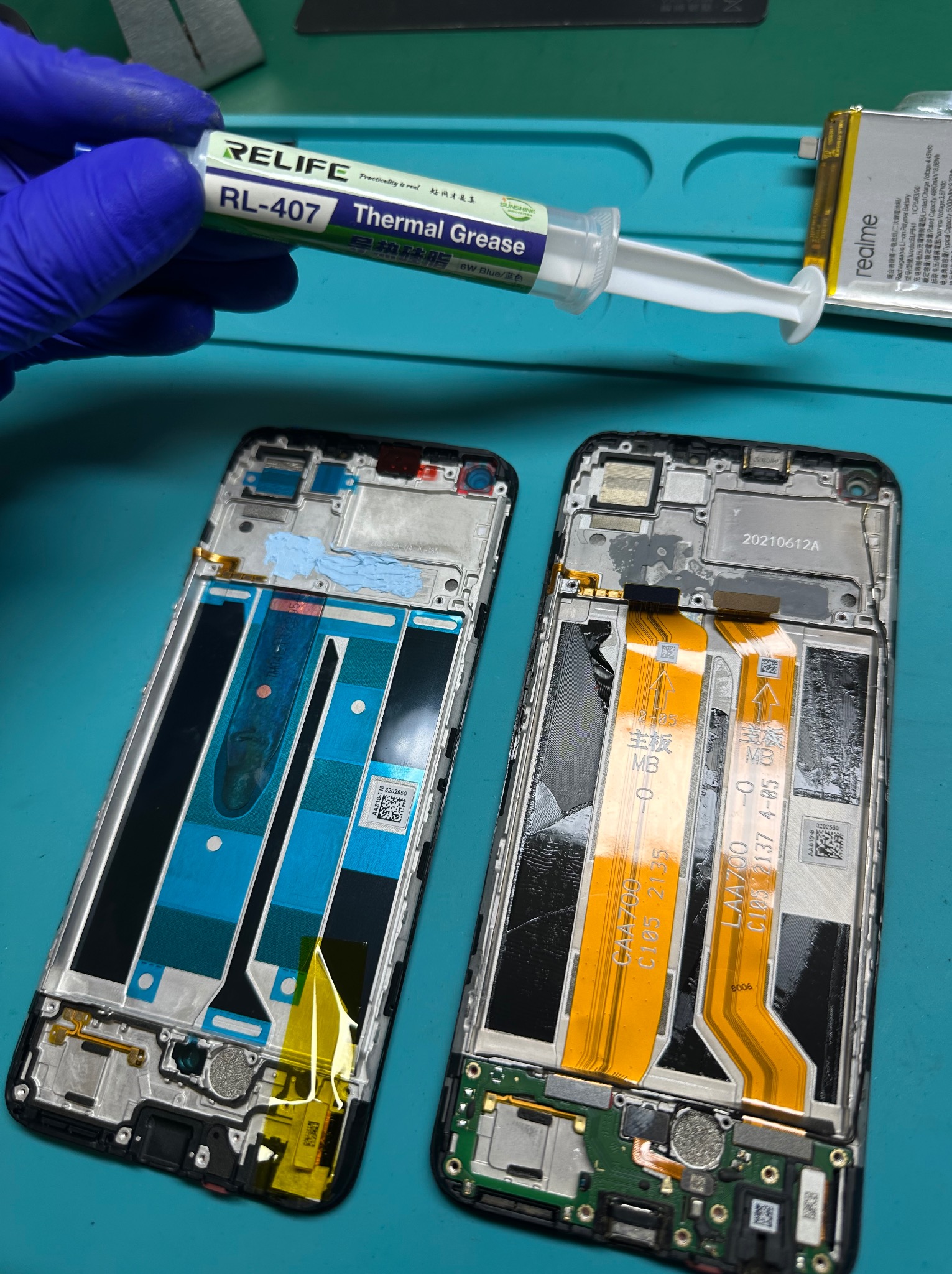 Dwa rozebrane smartfony obok siebie na zielonej macie serwisowej; jeden z nałożoną pastą termoprzewodzącą, drugi z widocznymi taśmami i elementami elektroniki; w górnej części dłoń w niebieskiej...