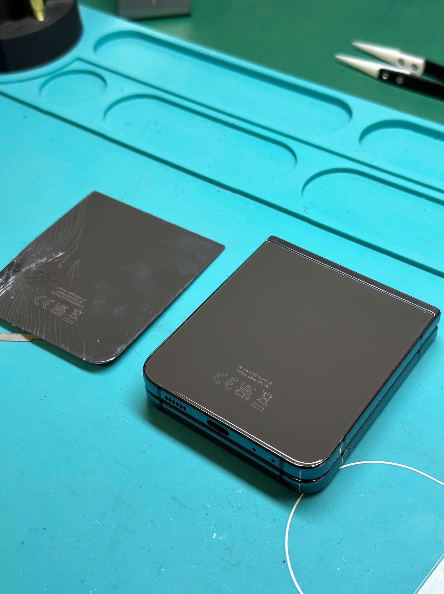 Uszkodzona tylna obudowa składanego smartfona leży obok nowego, sprawnego elementu na niebieskiej macie serwisowej z narzędziami w tle.