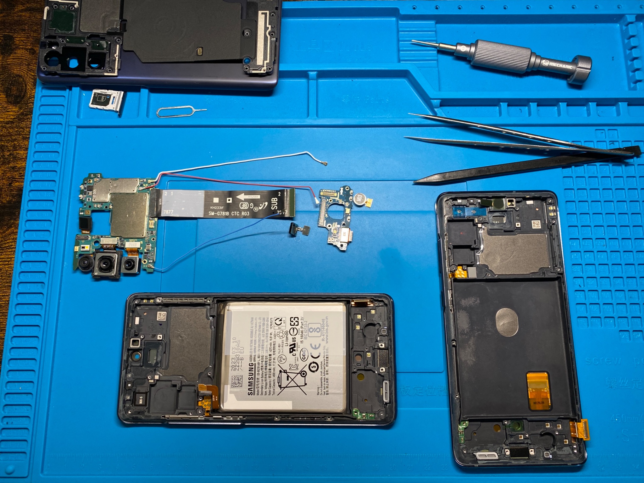 Rozłożony smartfon na niebieskiej macie serwisowej, widoczne narzędzia: śrubokręt, pęsety, obudowa, płyta główna, bateria i drobne elementy.