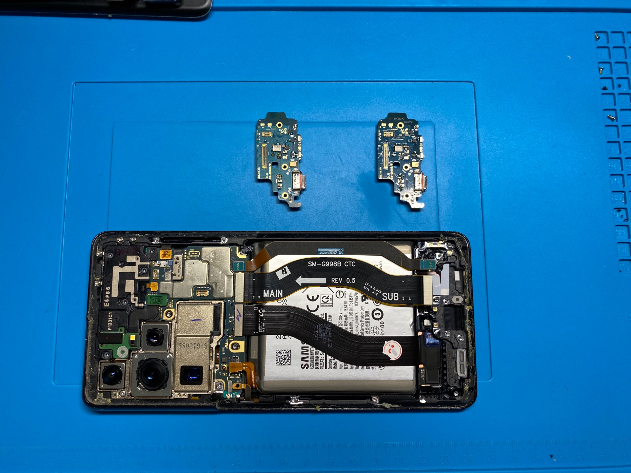Rozłożony smartfon Samsung na niebieskiej macie serwisowej, widoczne wewnętrzne komponenty, w tym bateria, płyta główna i dwie odłączone mniejsze płytki.