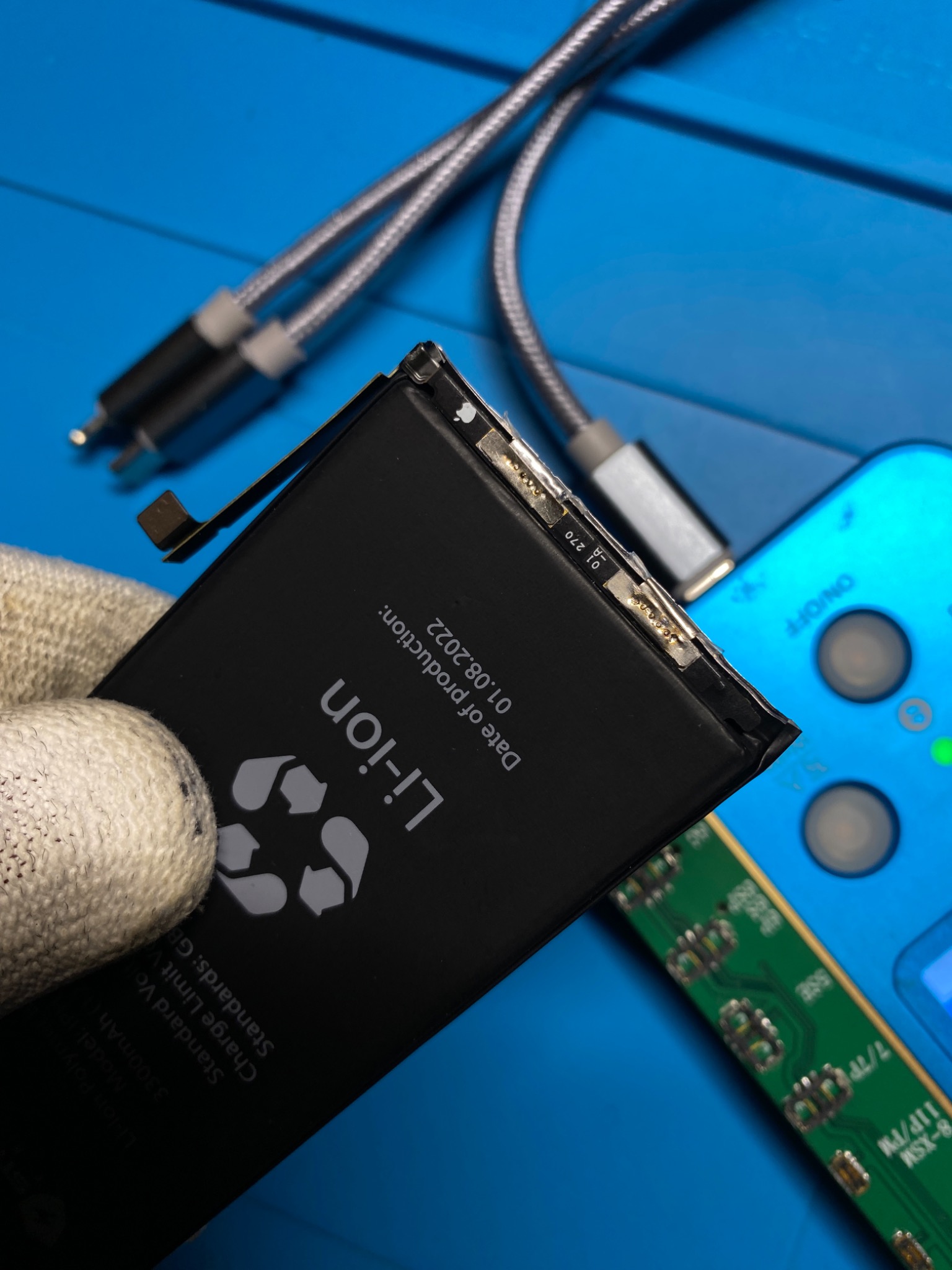 Wymiana baterii litowo-jonowej w telefonie; bateria w rękawicy ochronnej na tle niebieskiej maty serwisowej, widoczny kabel USB i fragment płytki PCB.
