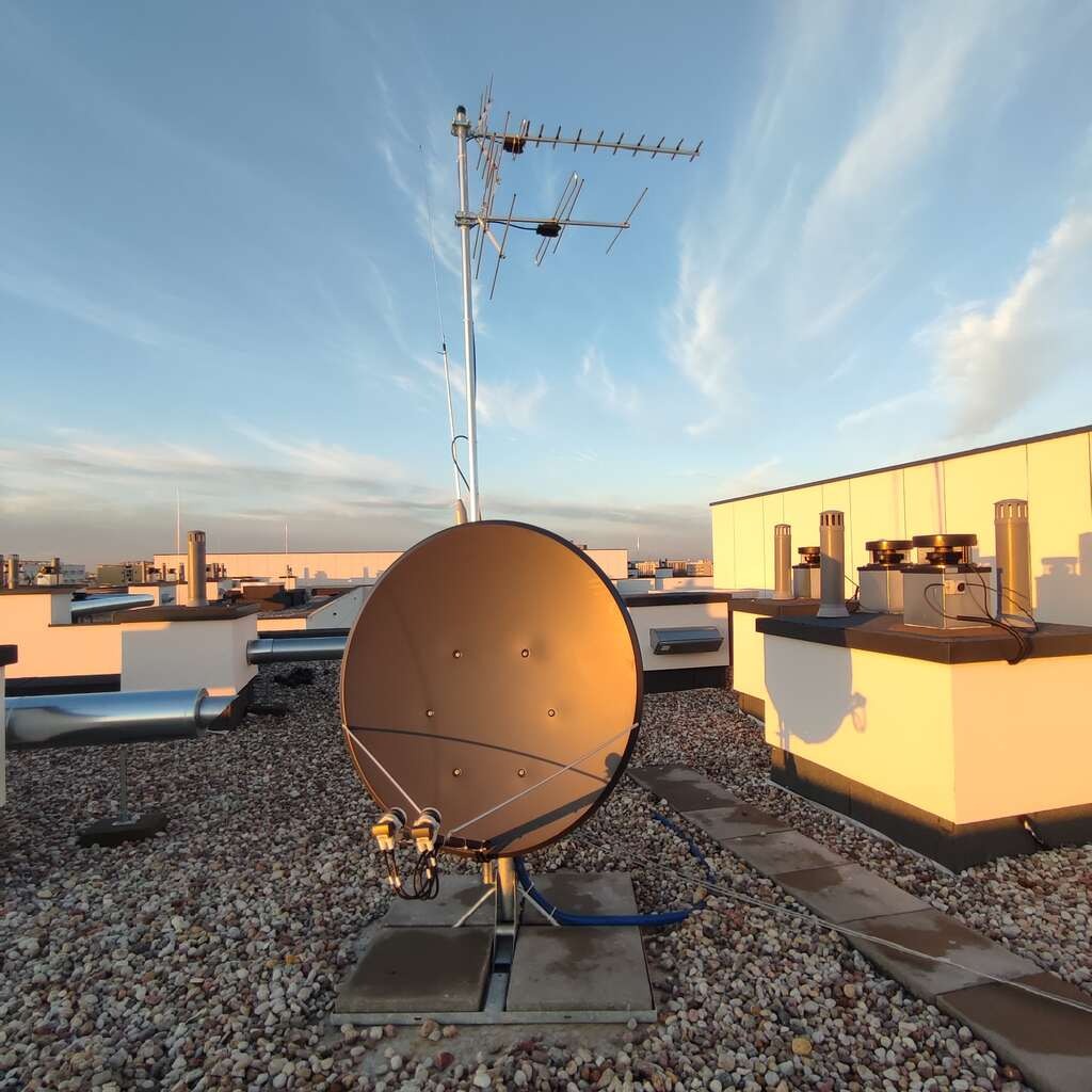 Antena satelitarna i antena naziemna zamontowane na płaskim dachu budynku z widocznymi elementami wentylacji i niebem w tle.