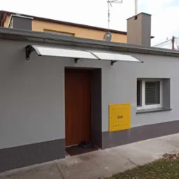 Szara elewacja budynku z brązowymi drzwiami wejściowymi pod białą, łukową markizą, ż&oacute;łta skrzynka gazowa z napisem 'GAZ' obok drzwi, białe okno z żaluzjami, ciemnoszara podmur&oacute;wka.