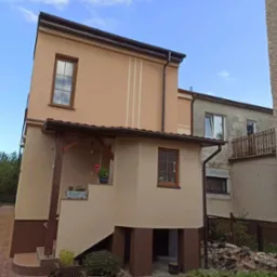 Dwukondygnacyjny dom po ociepleniu i pomalowaniu elewacji na beżowo, z brązowymi oknami PCV i ozdobnymi pionowymi paskami, widoczne kwiaty w donicach na schodach wejściowych, w tle budynek przed...