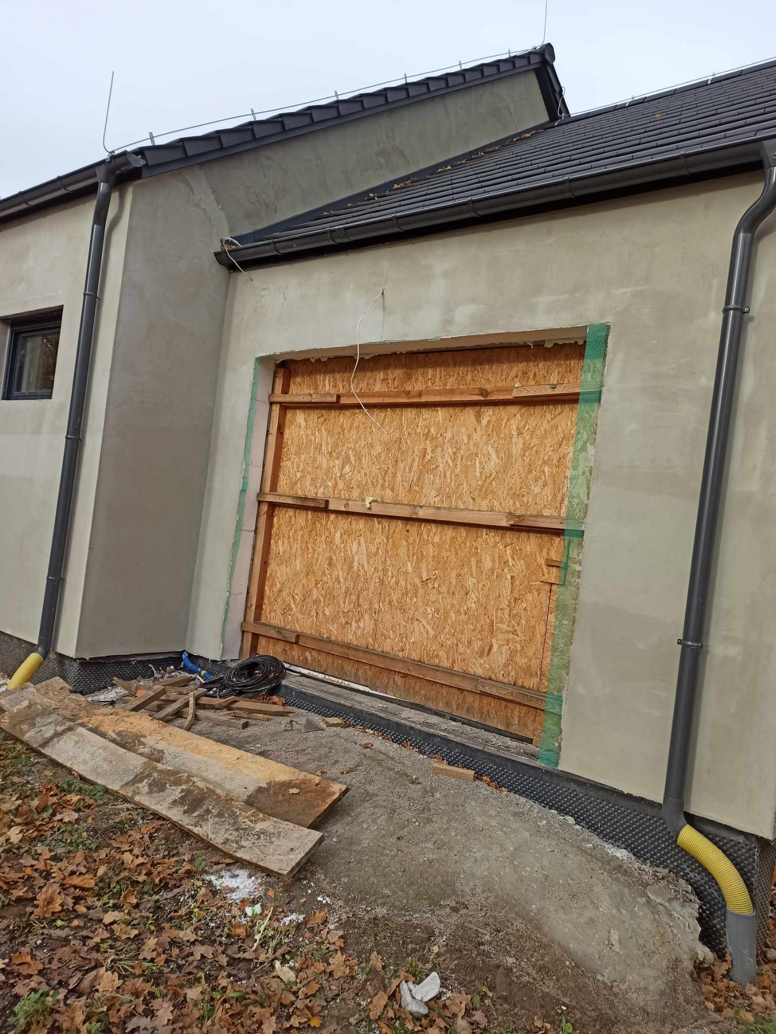 Budynek w trakcie prac elewacyjnych: ściana otynkowana, okno zabite płytą OSB, widoczne elementy ocieplenia, rynna i liście na ziemi.