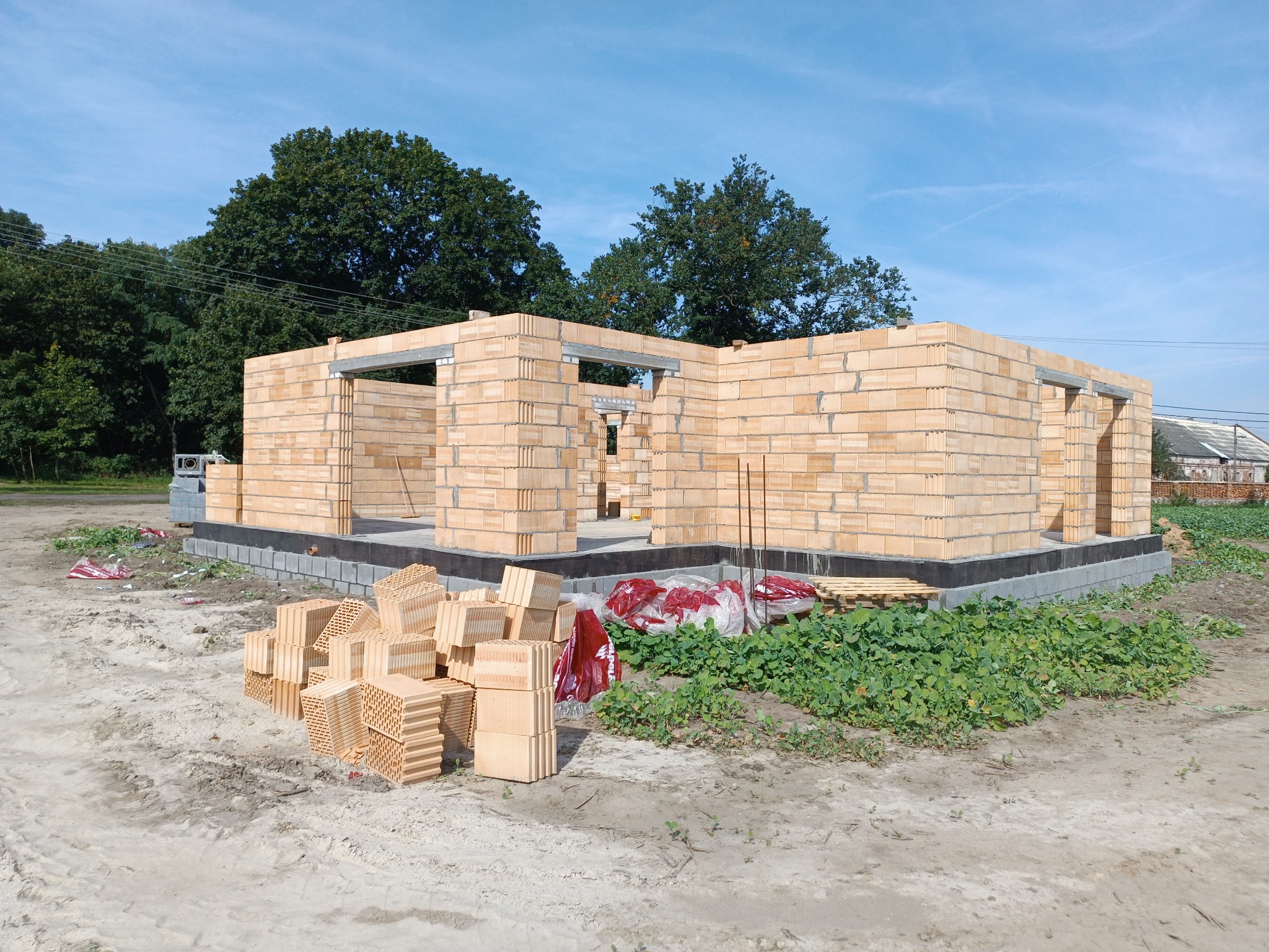 Wznoszenie ścian parterowego domu z bloczków ceramicznych na fundamencie z izolacją, widoczne nadproża okienne i drzwiowe, stos bloczków na pierwszym planie, tło: drzewa i błękitne niebo.