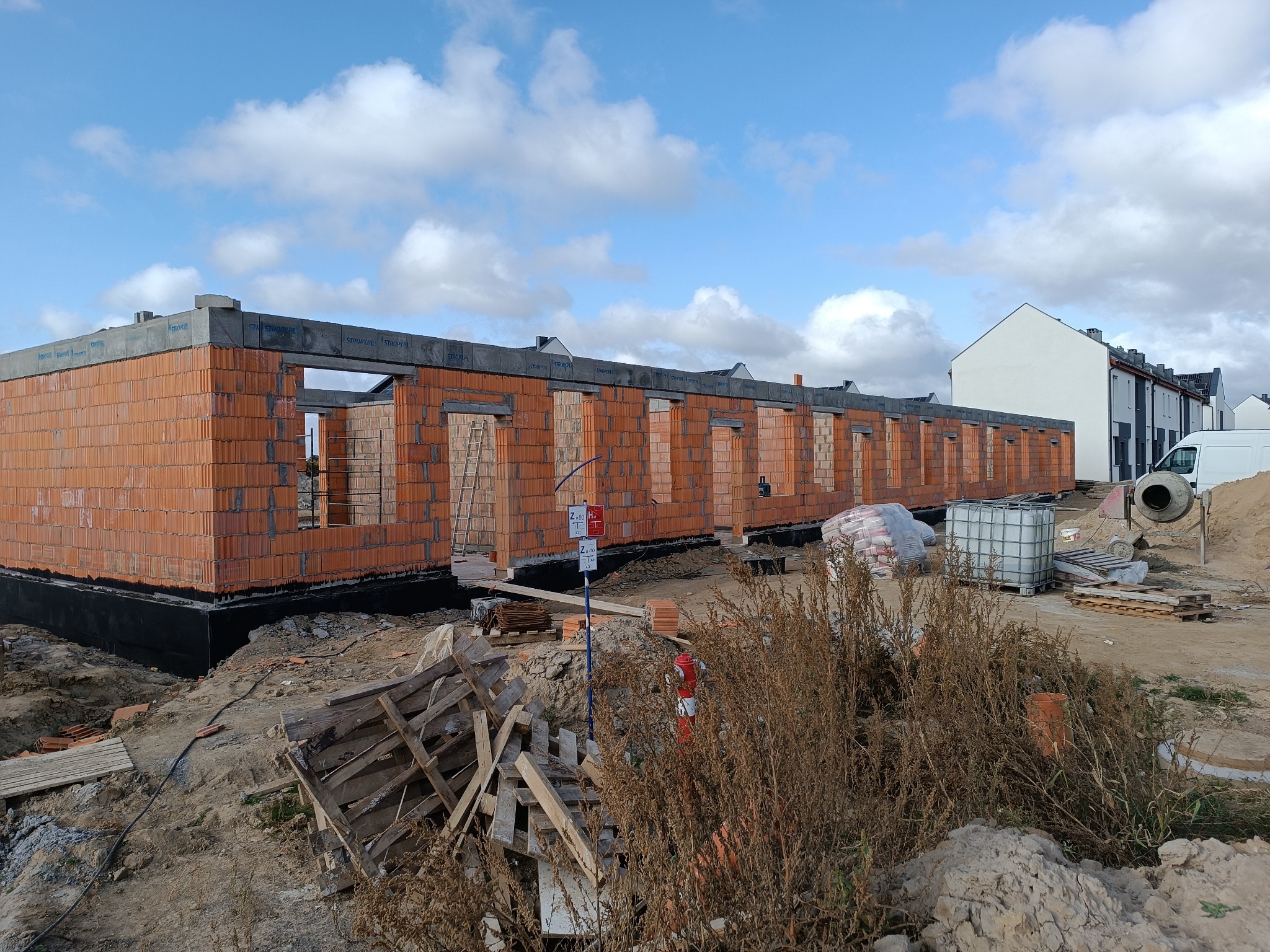 Wznoszenie ścian parterowego domu z czerwonej cegły, widoczne fundamenty z czarną izolacją, palety, worki z zaprawą i betoniarka na placu budowy.