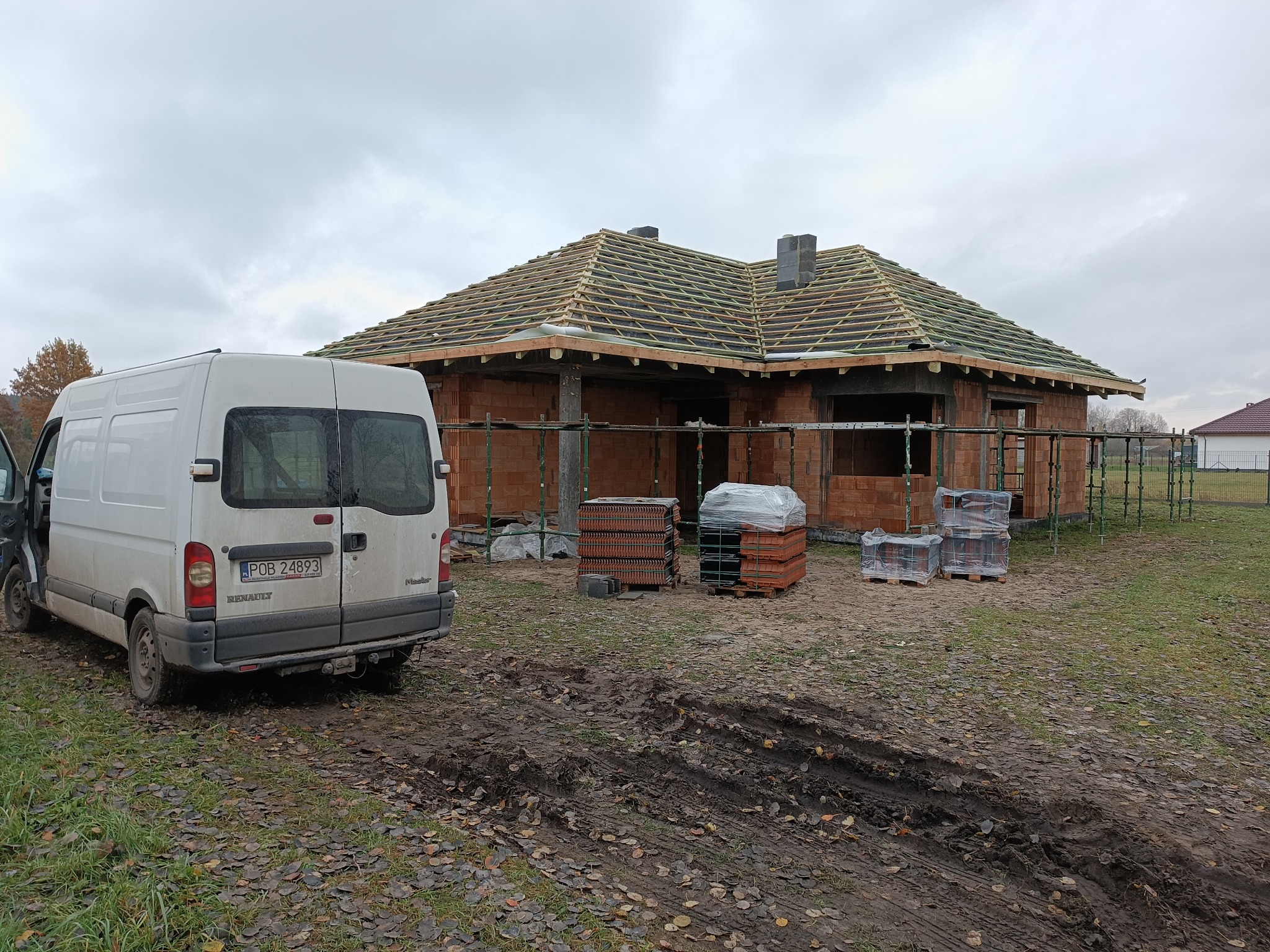Wznoszony budynek z czerwonej cegły z widoczną konstrukcją dachu, częściowo pokrytą papą, na tle pochmurnego nieba; obok zaparkowany biały bus i palety z dachówką.