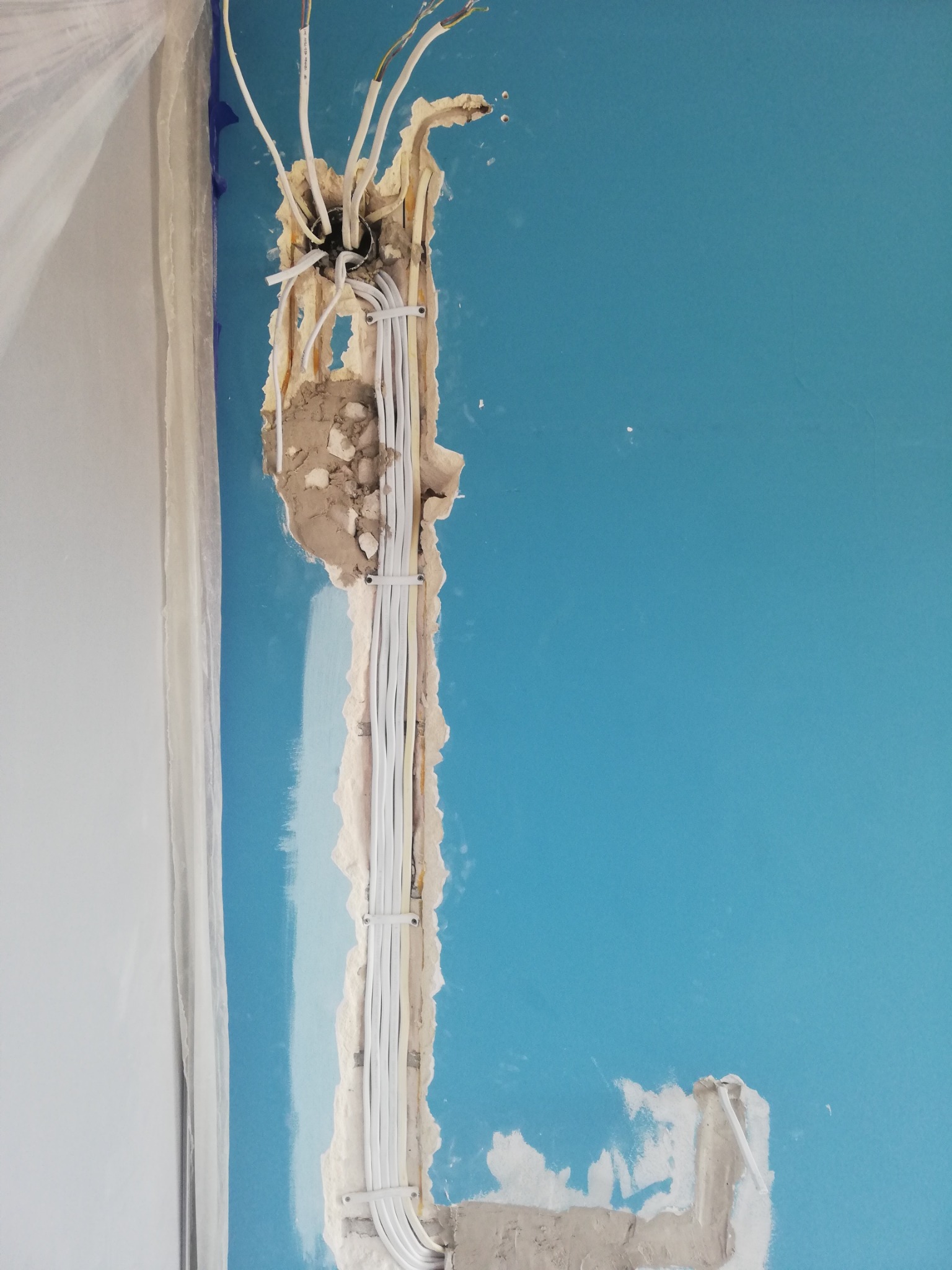 Instalacja elektryczna w ścianie o błękitnym kolorze, widoczne przewody prowadzone w bruździe, zabezpieczone uchwytami, świeżo po położeniu gładzi szpachlowej.