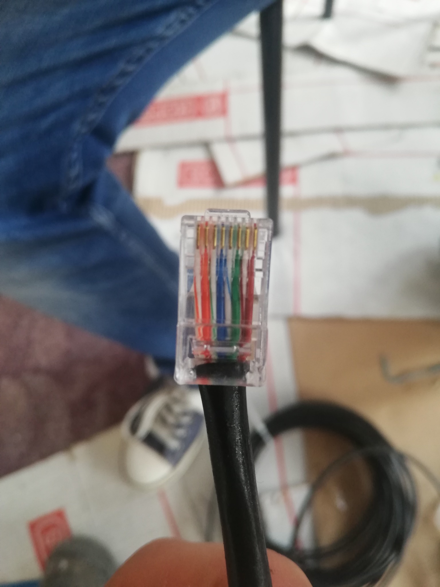 Złącze RJ45 z widocznymi kolorowymi przewodami, czarny kabel, w tle fragment podłogi z kartonami i fragment buta.