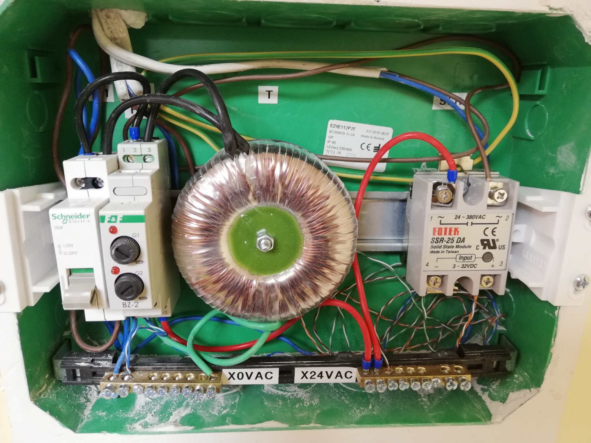 Wnętrze zielonej skrzynki elektrycznej z transformatorem toroidalnym, dwoma przekaźnikami, listwami zaciskowymi i okablowaniem, widok z góry.