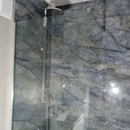 Nowoczesna kabina prysznicowa z deszczownicą i szklanymi drzwiami, wykończona płytami imitującymi marmur w odcieniach szarości i błękitu, z widocznym kratką wentylacyjną.