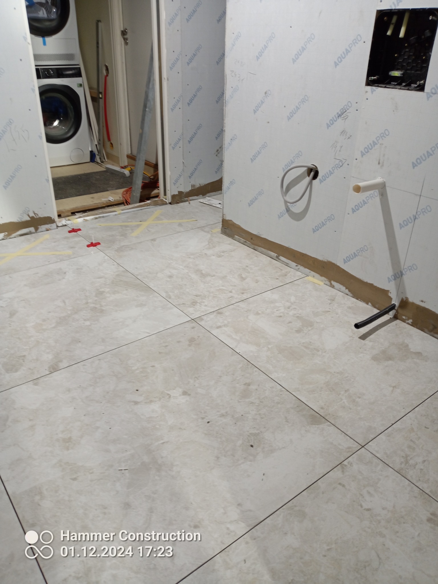 Łazienka w trakcie remontu: ułożone płytki podłogowe w odcieniach beżu z widocznymi krzyżykami dystansowymi, ściany wyłożone płytami Aquapro, widoczne instalacje hydrauliczne i elektryczne, pralka...