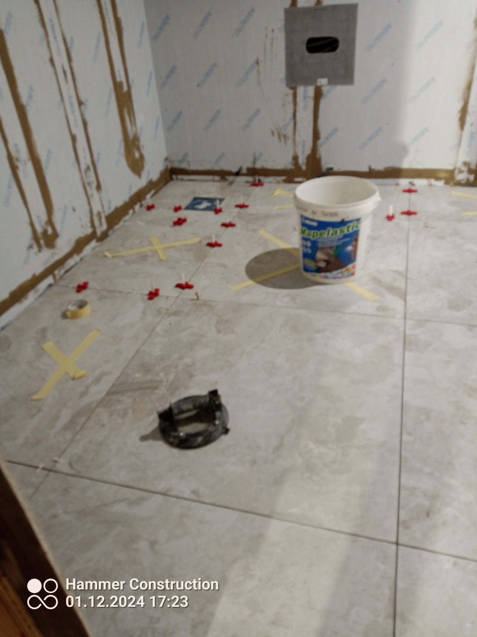 Podczas układania płytek podłogowych: widoczny biały pojemnik z klejem Mapei, krzyżyki dystansowe, żółta taśma maskująca i narzędzie do cięcia płytek.