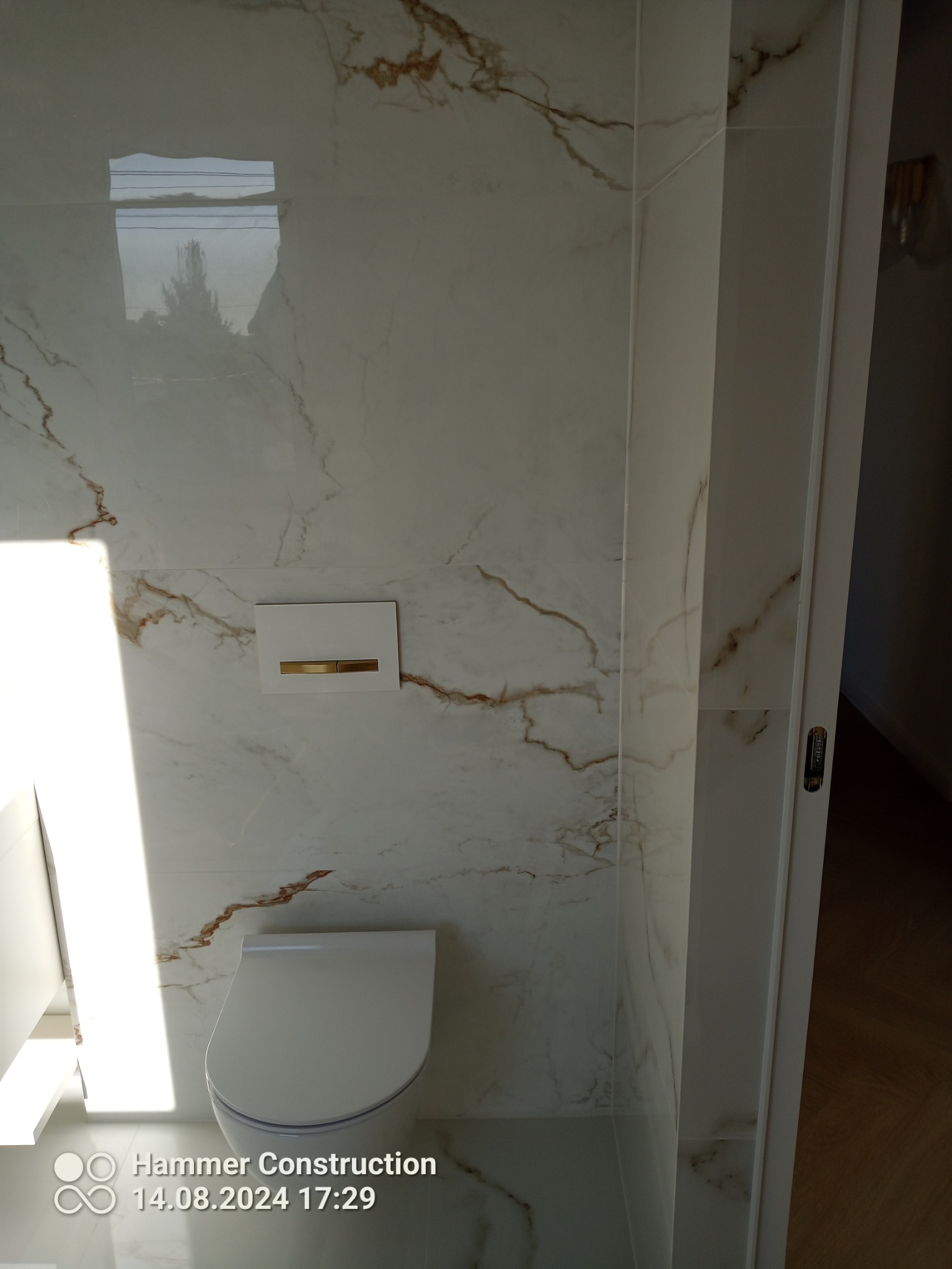 Nowoczesna toaleta podwieszana z białym panelem spłukującym ze złotym akcentem, na ścianie płytki imitujące marmur z brązowymi żyłami, widoczne odbicie drzewa w płytkach.