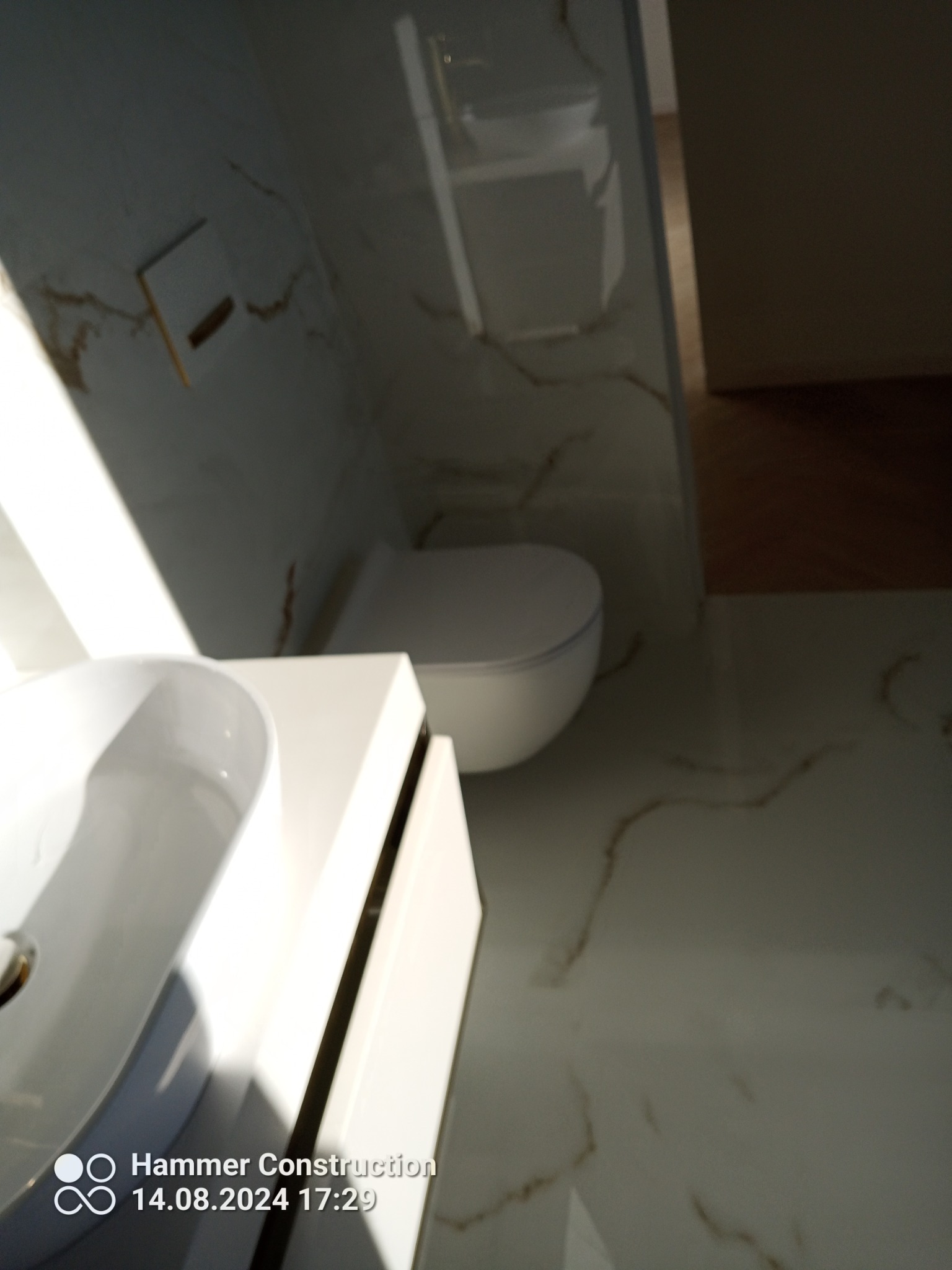 Nowoczesna łazienka z białymi płytkami imitującymi marmur, wisząca toaleta i umywalka z szafką w minimalistycznym stylu.