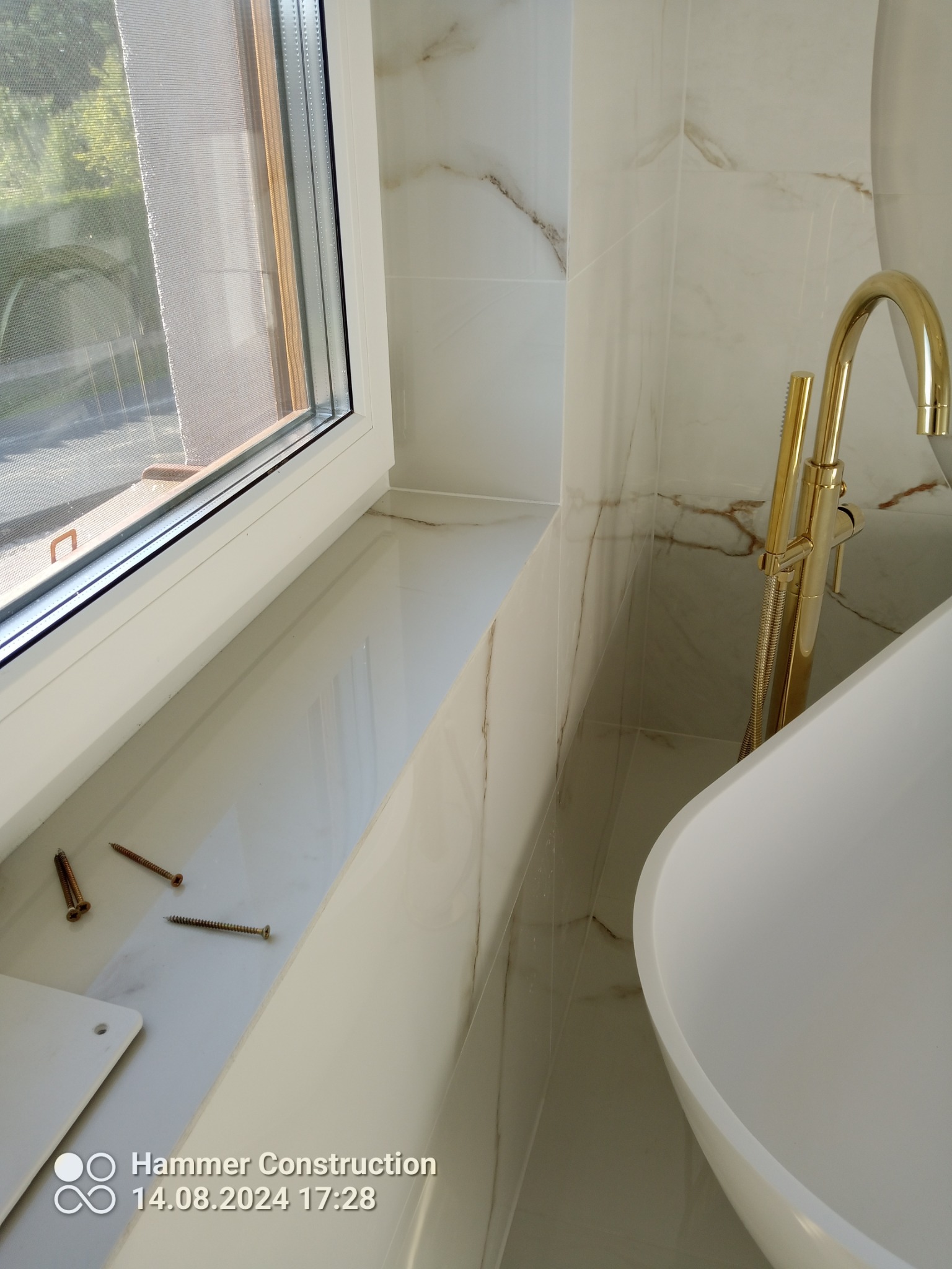 Biała umywalka wolnostojąca z złotą baterią na tle marmurowych płytek, widok na okno z trzema śrubami na parapecie.