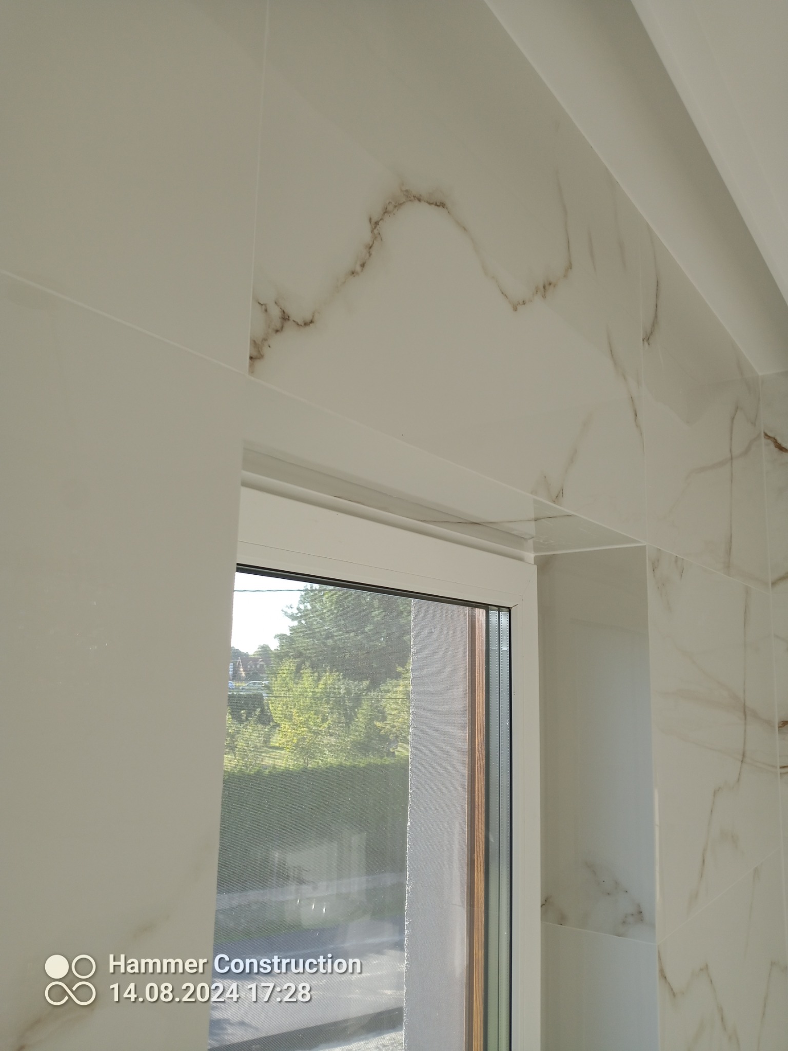 Jasne, marmurowe płytki ścienne z brązowymi żyłkami, ułożone wokół okna z widokiem na zieleń.