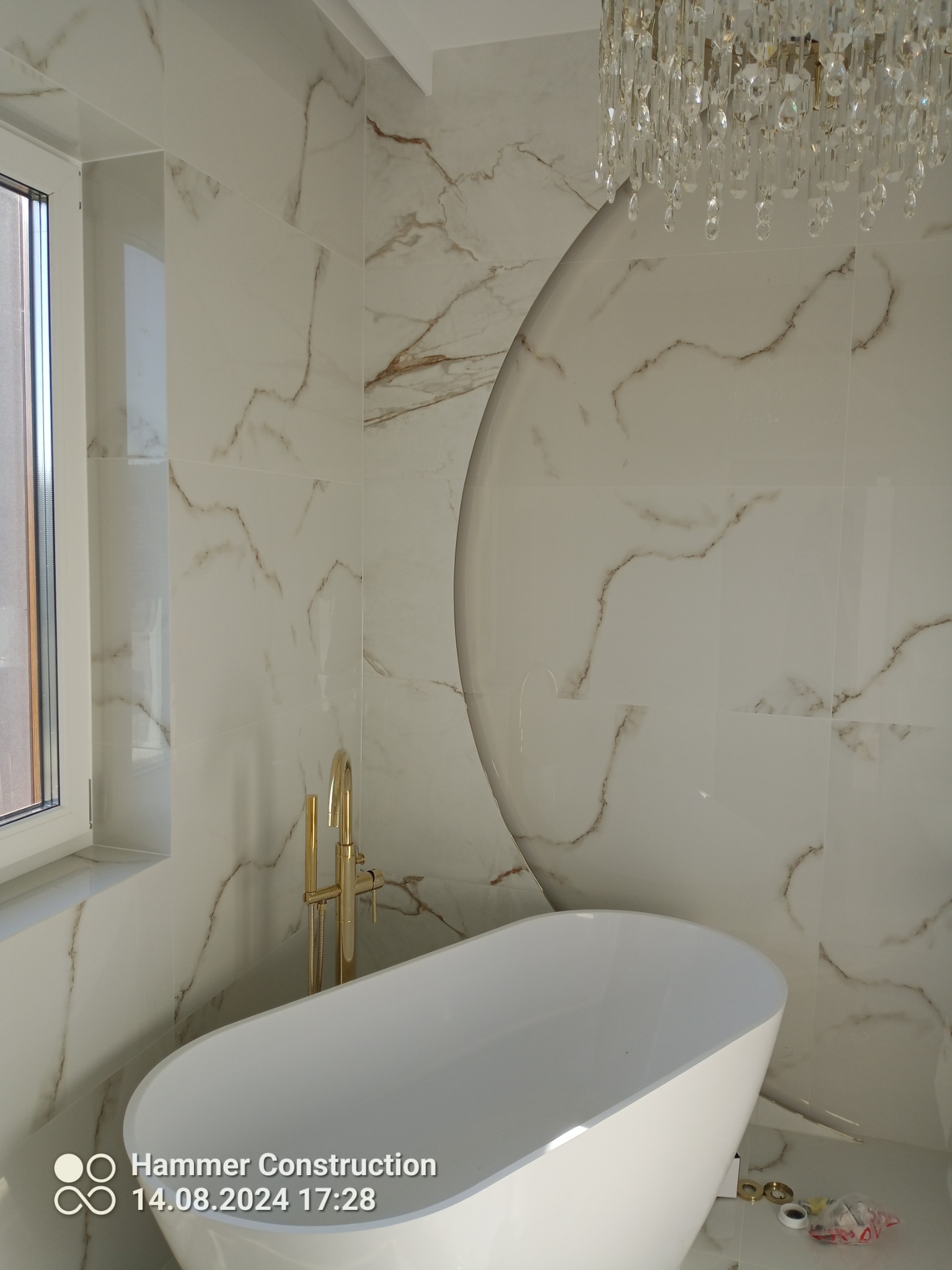 Elegancka łazienka z wanną wolnostojącą, złotą armaturą i marmurowymi płytkami ściennymi, doświetlona kryształowym żyrandolem.