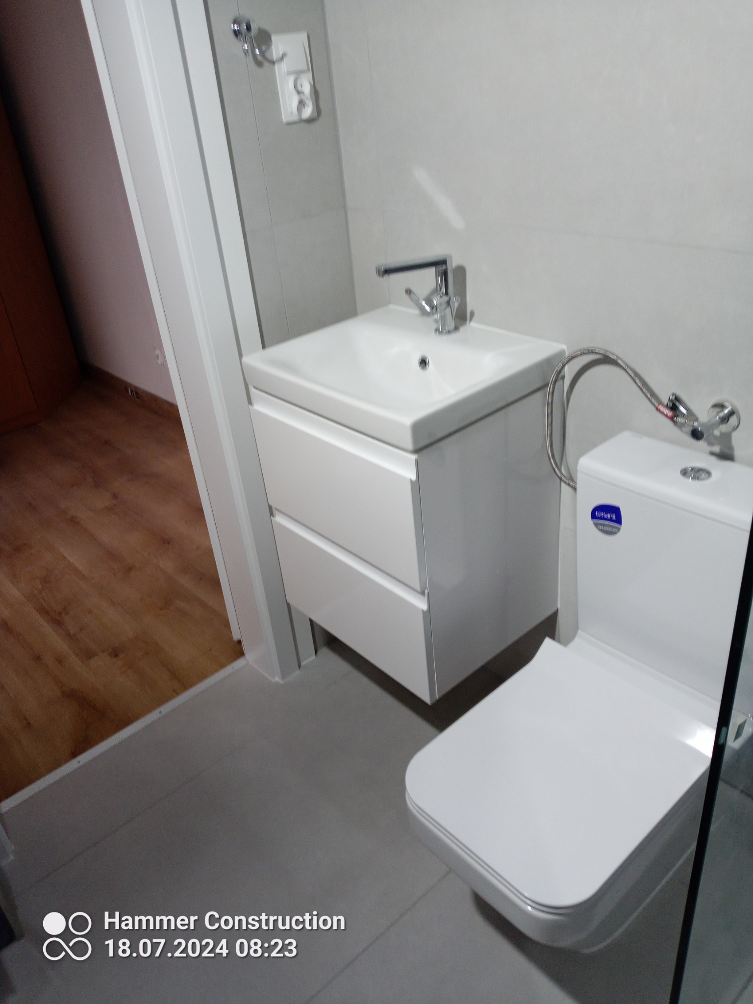 Nowoczesna, jasna łazienka z białym zestawem: umywalka z szafką i wisząca toaleta z bidetką, szare płytki na ścianach i podłodze, widoczny fragment drewnianej podłogi w sąsiednim pomieszczeniu.