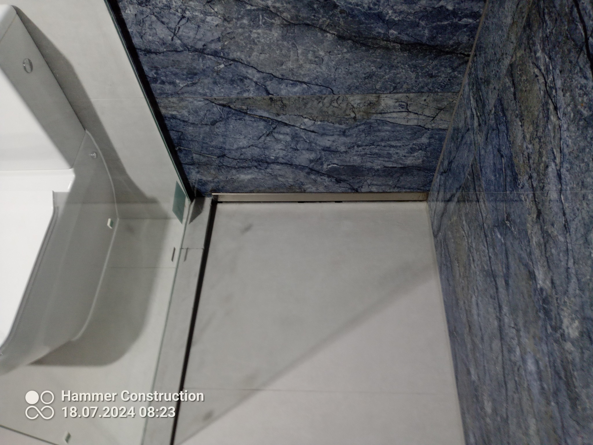 Fragment łazienki z kabiną prysznicową i niebieskimi płytkami imitującymi marmur na suficie i ścianie, widoczne mocowania szklanej kabiny.