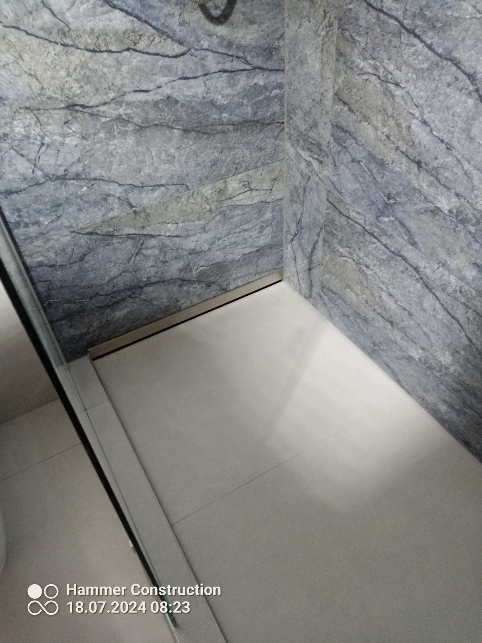 Wykończony narożnik kabiny prysznicowej z szarymi płytkami imitującymi marmur na ścianach i jasnymi płytkami na podłodze, widoczny profil kabiny.