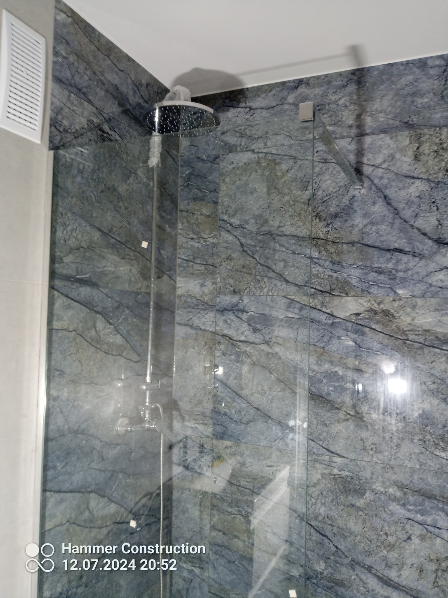 Nowoczesna kabina prysznicowa z deszczownicą i szklanymi drzwiami, wykończona płytami imitującymi marmur w odcieniach szarości i błękitu, z widocznym kratką wentylacyjną.