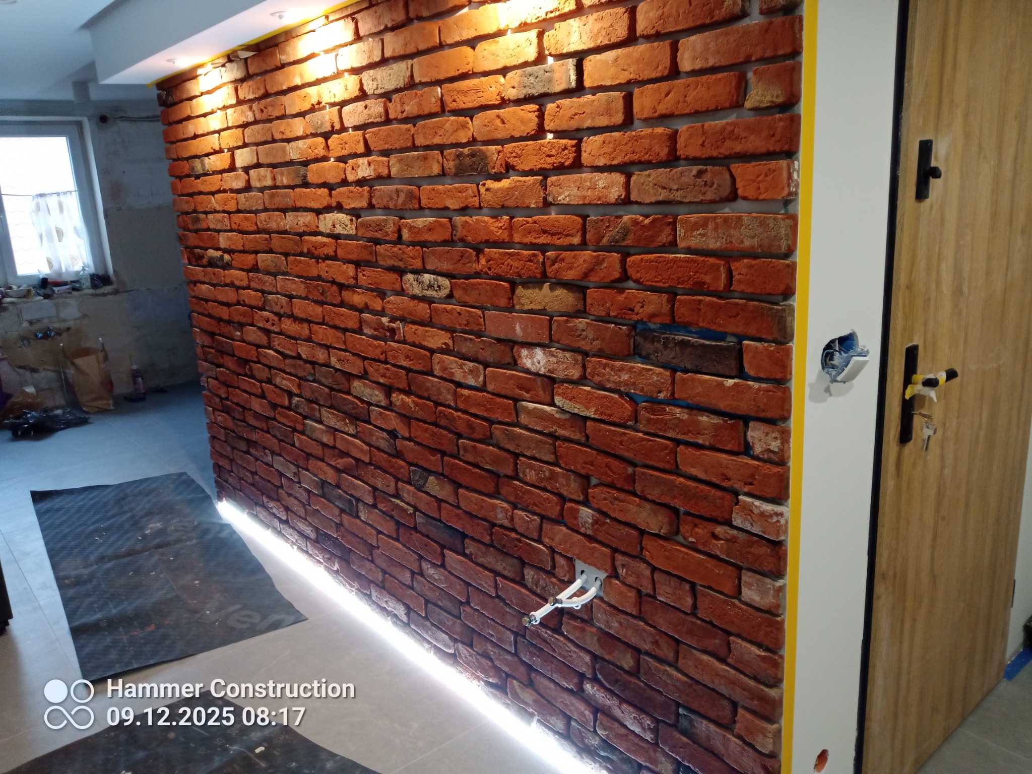 Ściana z cegły klinkierowej z podświetleniem LED u dołu, widoczna instalacja elektryczna, fragment drzwi. Wykończenie wnętrza w surowym stylu.