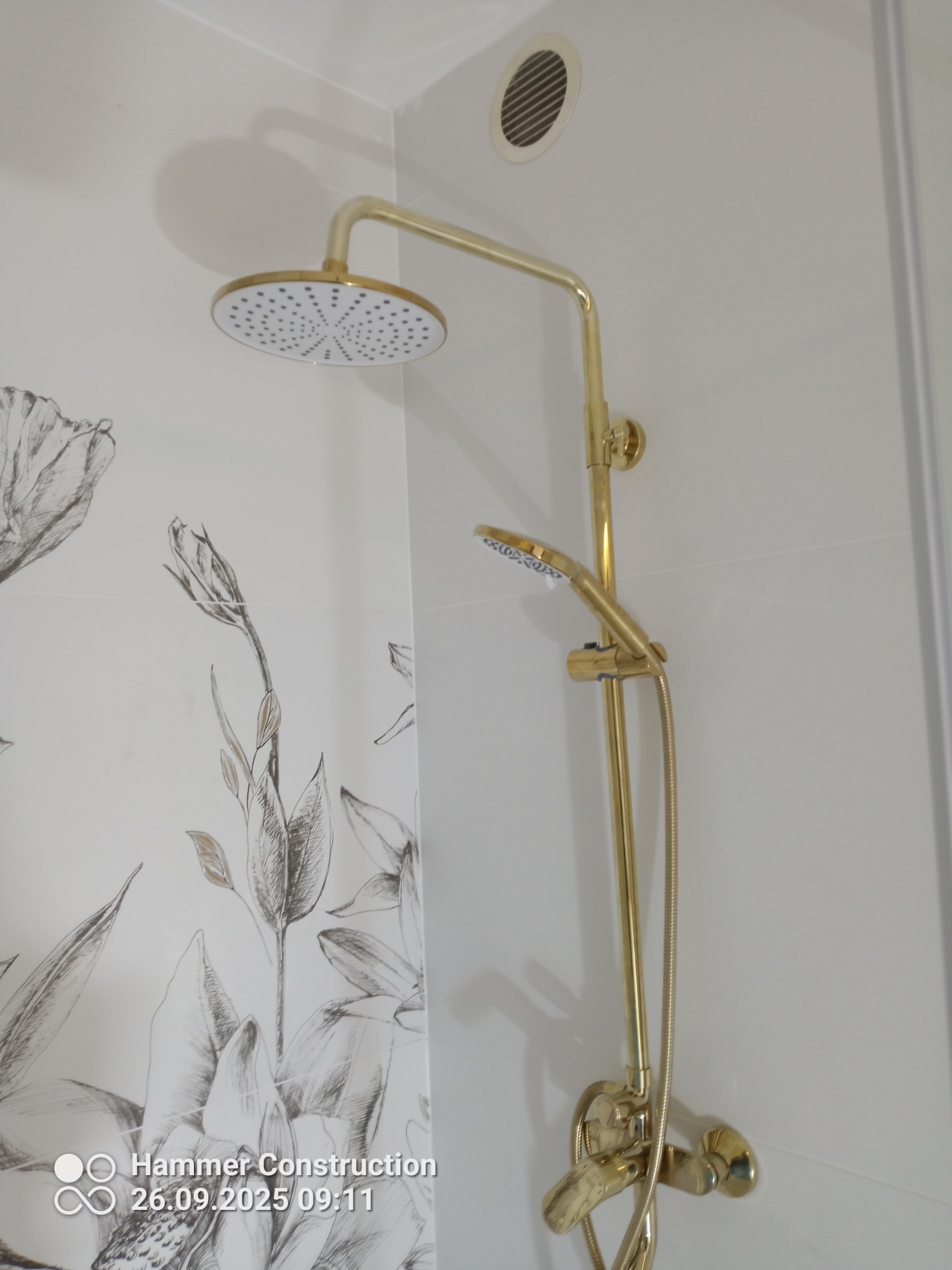 Bateria prysznicowa w odcieniach złota na tle białych płytek z motywem kwiatowym. Widoczny również fragment wentylacji i kabiny prysznicowej.
