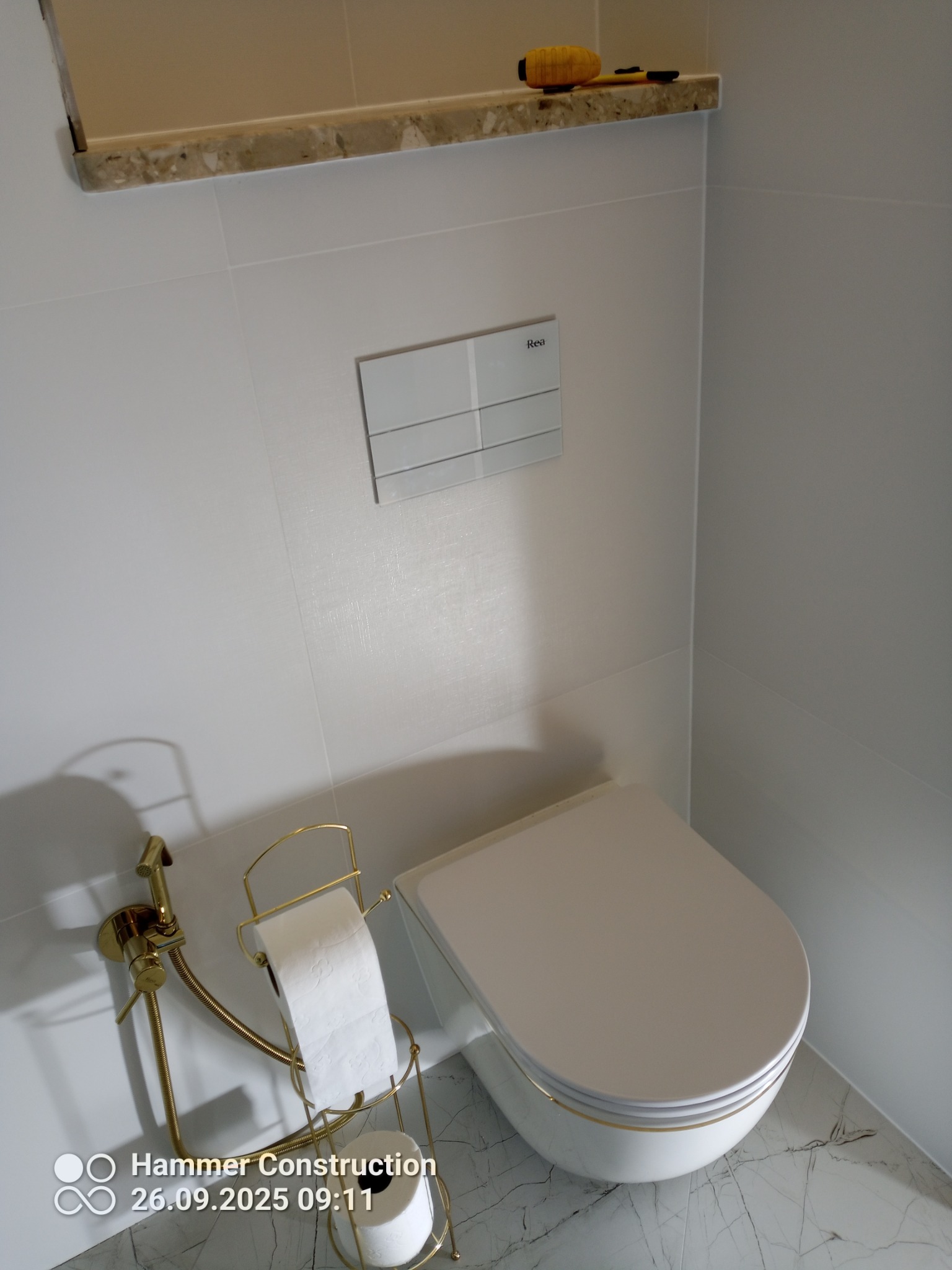 Nowoczesna toaleta podwieszana z białymi płytkami i marmurową podłogą. Widoczny przycisk spłukujący i złoty uchwyt na papier toaletowy z bidetką.