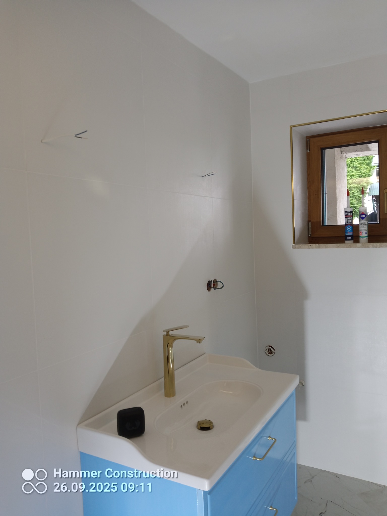 Jasna łazienka po remoncie: białe płytki, błękitna szafka pod umywalką z złotą armaturą. W oknie widoczne silikony. Minimalistyczne wieszaki na ścianie.