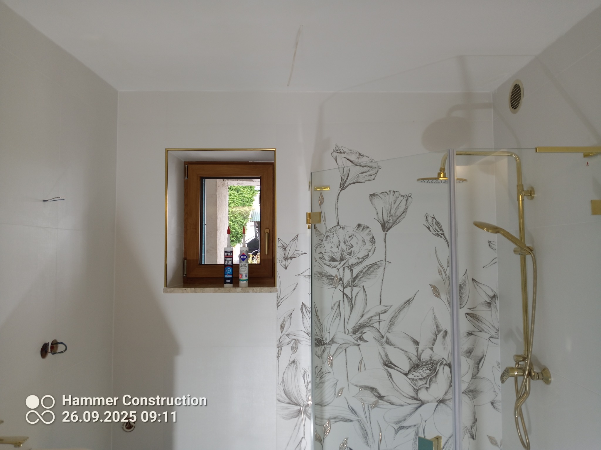 Wykończona łazienka z kabiną prysznicową ozdobioną motywem kwiatowym, złotą armaturą i oknem z widocznymi produktami do montażu.