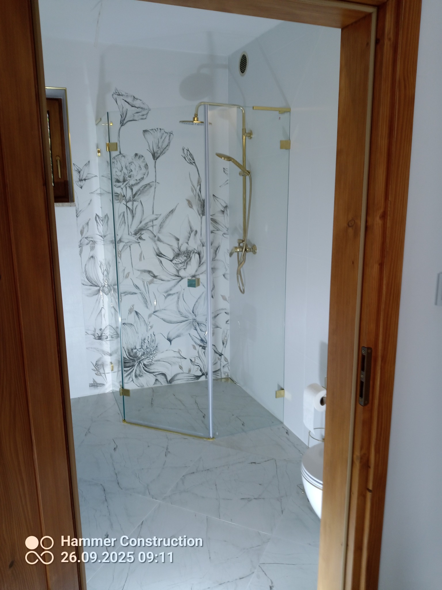 Nowoczesna kabina prysznicowa z złotymi detalami i wzorem kwiatowym na ścianie, widoczna zza drewnianej futryny drzwi. Marmurowe płytki podłogowe.