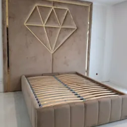 Beżowe łóżko tapicerowane z wysokim wezgłowiem zdobionym złotym geometrycznym wzorem diamentu, widoczny stelaż z drewnianych listew, białe ściany w tle.