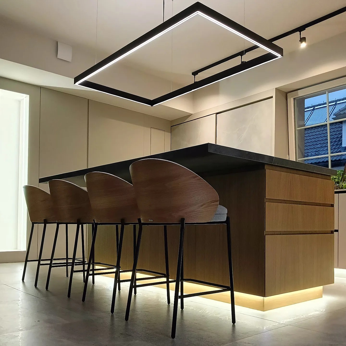 Nowoczesna wyspa kuchenna z drewnianymi frontami i czarnym blatem, oświetlona prostokątną lampą LED i podświetleniem cokołu, z czterema drewnianymi hokerami z czarnymi nogami.