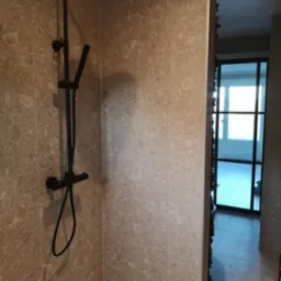 Nowoczesna kabina prysznicowa z czarną armaturą na tle beżowych płytek imitujących kamień, widoczny pędzel na podłodze i fragment czarnej ramy drzwi w tle.