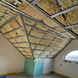 Poddasze w trakcie izolacji: wełna mineralna w metalowym stelażu, widoczna drewniana belka, okrągłe okno i fragment płyty gipsowo-kartonowej.